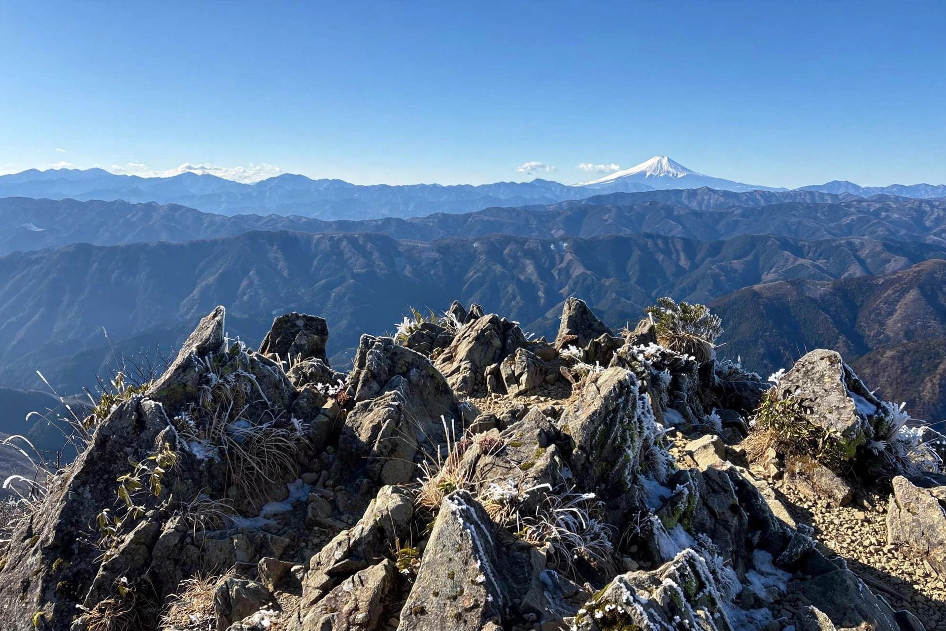 Snow-capped Mt. Fuji rising above layered blue mountain ridges, viewed past jagged rocky summit outcrops on a clear winter morning